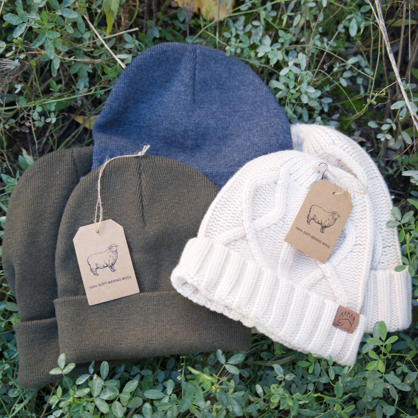 Assorted 100% Soft Merino Wool Beanies in cream, olive, and charcoal displayed on green foliage. Stylish and warm for outdoor wear.