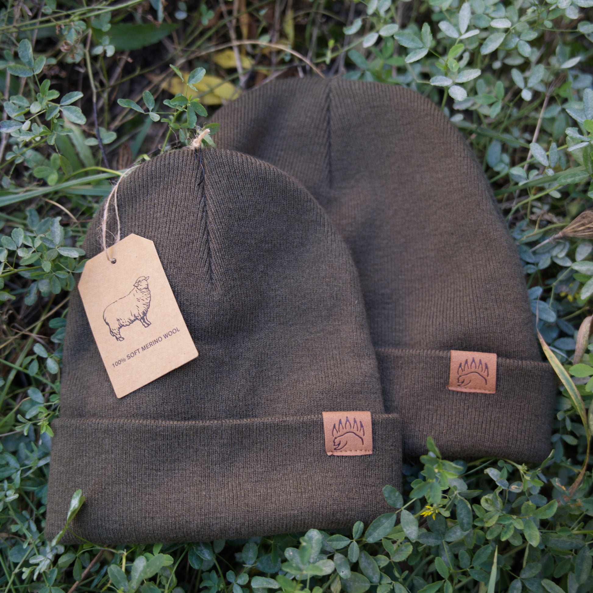 Pair of olive 100% Soft Merino Wool Beanies with tags and bear paw logos, laid on grass for natural product presentation.