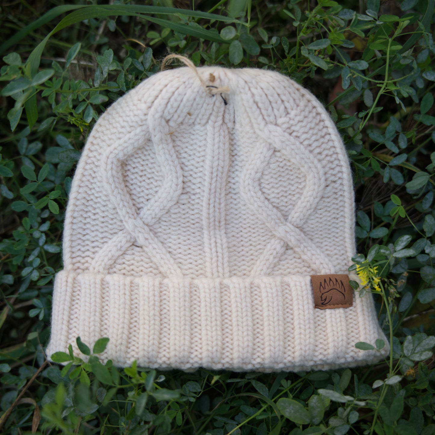 Single cream cable-knit 100% Soft Merino Wool Beanie with ribbed cuff and bear paw patch, placed on green foliage.