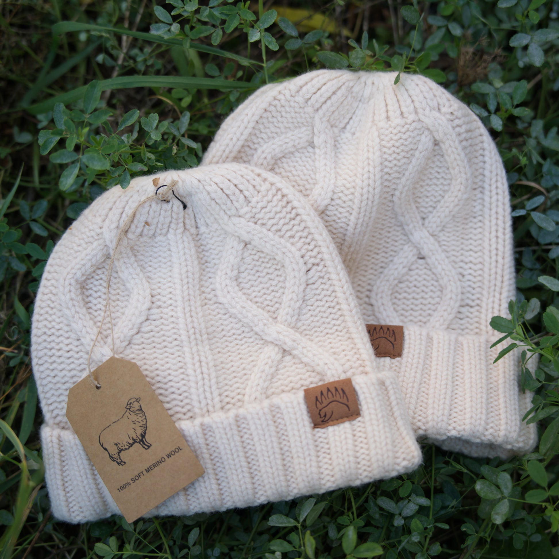 Two cream cable-knit 100% Soft Merino Wool Beanies with ribbed cuffs and bear paw patches, styled for winter fashion.