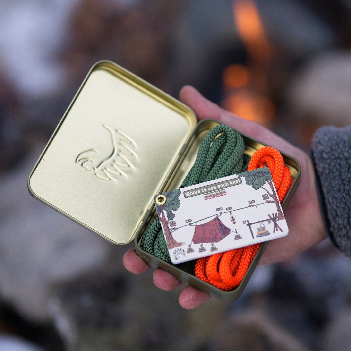 Hand holding The Bear Essentials Knot Tying Kit tin with ropes and knot cards, perfect for learning camping knots by the fire.
