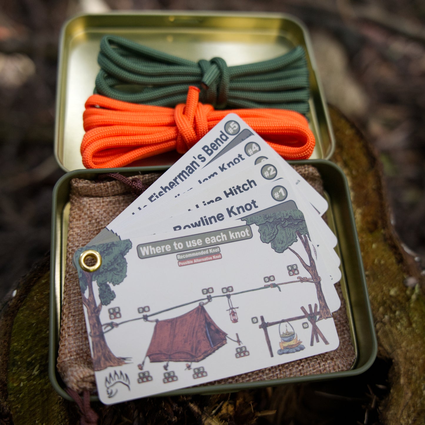Open tin of The Bear Essentials Knot Tying Kit showing ropes and illustrated knot cards, designed for learning top camping knots.