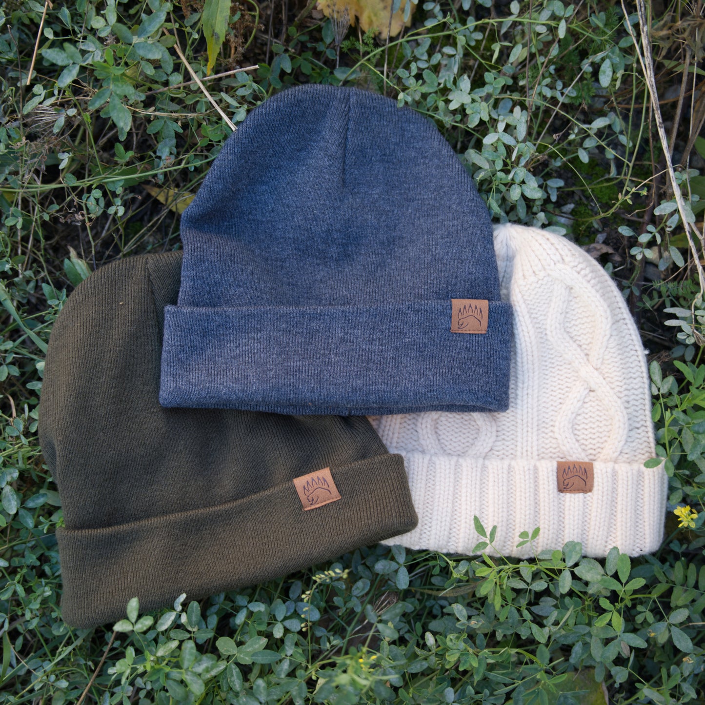 Three 100% Soft Merino Wool Beanies in cream, olive, and charcoal with bear paw logos, laid on grass for product display.