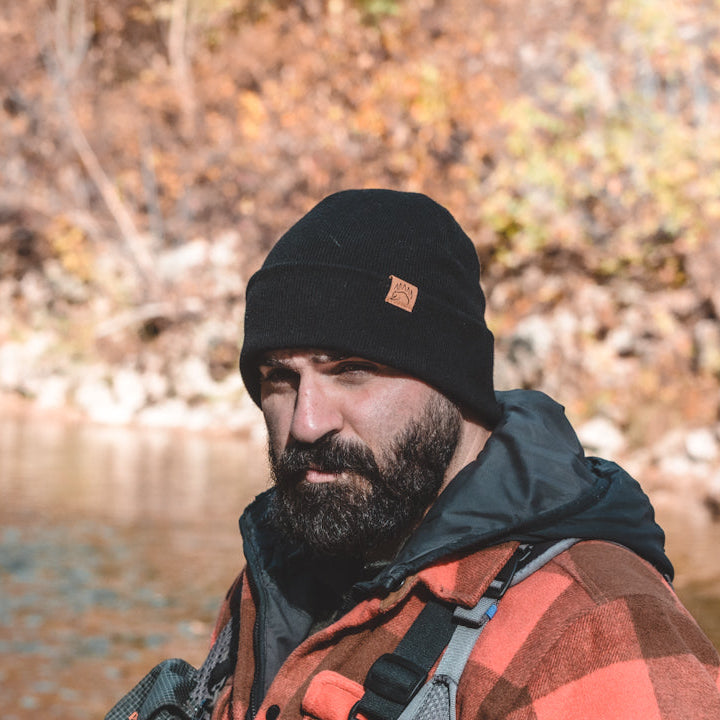 Man wearing a black beanie and plaid jacket standing by a body of water with autumn foliage.